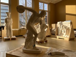 Im Vordergrund steht die Gipsfigur eines Diskuswerfers in einem hellen Raum, in dem auch andere Gipsfiguren gerade aus Boxen ausgepackt werden.