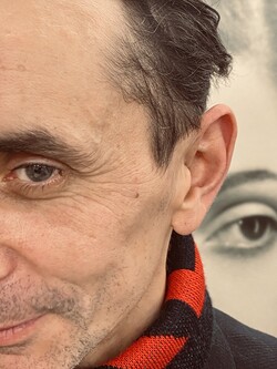 Nahaufnahme einer Gesichtshälfte eines Mannes mit schwarzem Haar und rot-schwarzem Schal. Im Hintergrund ein unscharfes schwarz-weiß Portrait einer Frau, deren Auge gut erkennbar ist.
