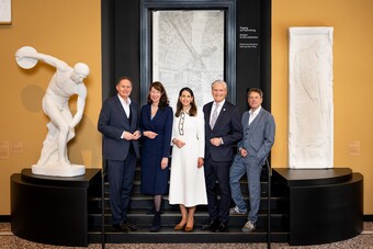 Vier Geschäftspersonen stehen vor goldener Wand neben weißer Skulptur in moderner Galerie