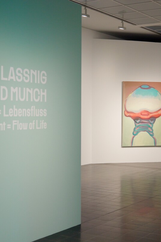 Galerieraum mit türkiser Wand, Text und zwei gerahmten Kunstwerken an weißer Wand