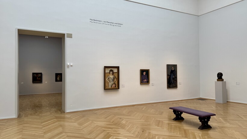 Heller Galerieraum mit Parkettboden, mehreren gerahmten Kunstwerken von Max Beckmann an weißen Wänden und einer Bank in der Mitte