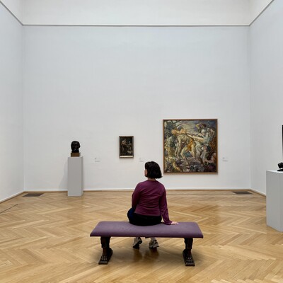 Person sitzt auf Bank in hellem Galerieraum mit Kunstwerken von Max Beckmann
