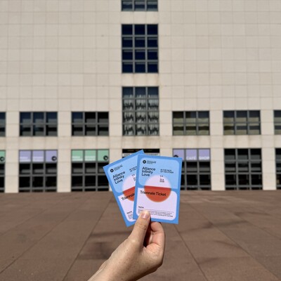 Hand hält zwei Tickets vor einem Gebäude mit vielen Fenstern