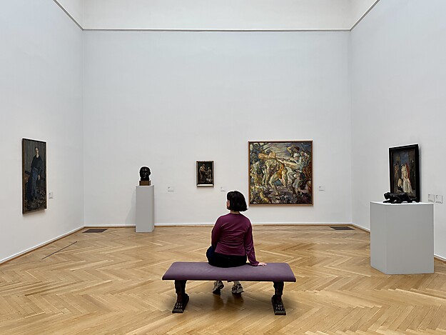 Person sitzt auf Bank in hellem Galerieraum mit Kunstwerken von Max Beckmann